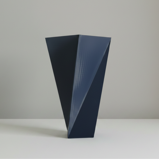 A modern geometric vase with a dark navy blue finish, featuring a faceted triangular design and subtle vertical ribbed texture, standing on a light gray surface against a neutral gray background.