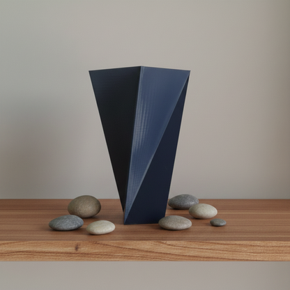 A dark navy blue geometric vase stands on a wooden surface with smooth stones around it against a plain beige wall.