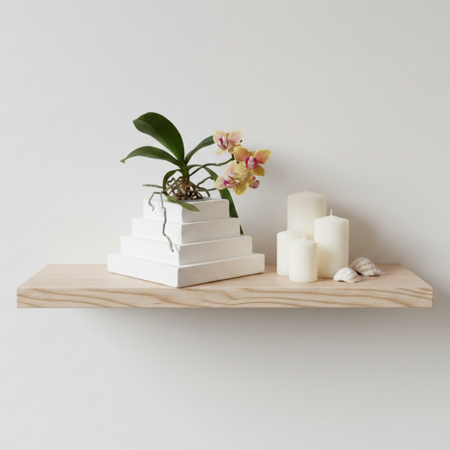 A light wood floating shelf mounted on a white wall, styled with a small potted orchid, white pyramid-shaped planter, and several white pillar candles with small shells.