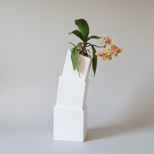 A white, tiered geometric planter made of stacked square blocks, holding a green leafy plant with yellow and pink orchid flowers, set against a plain light gray background.