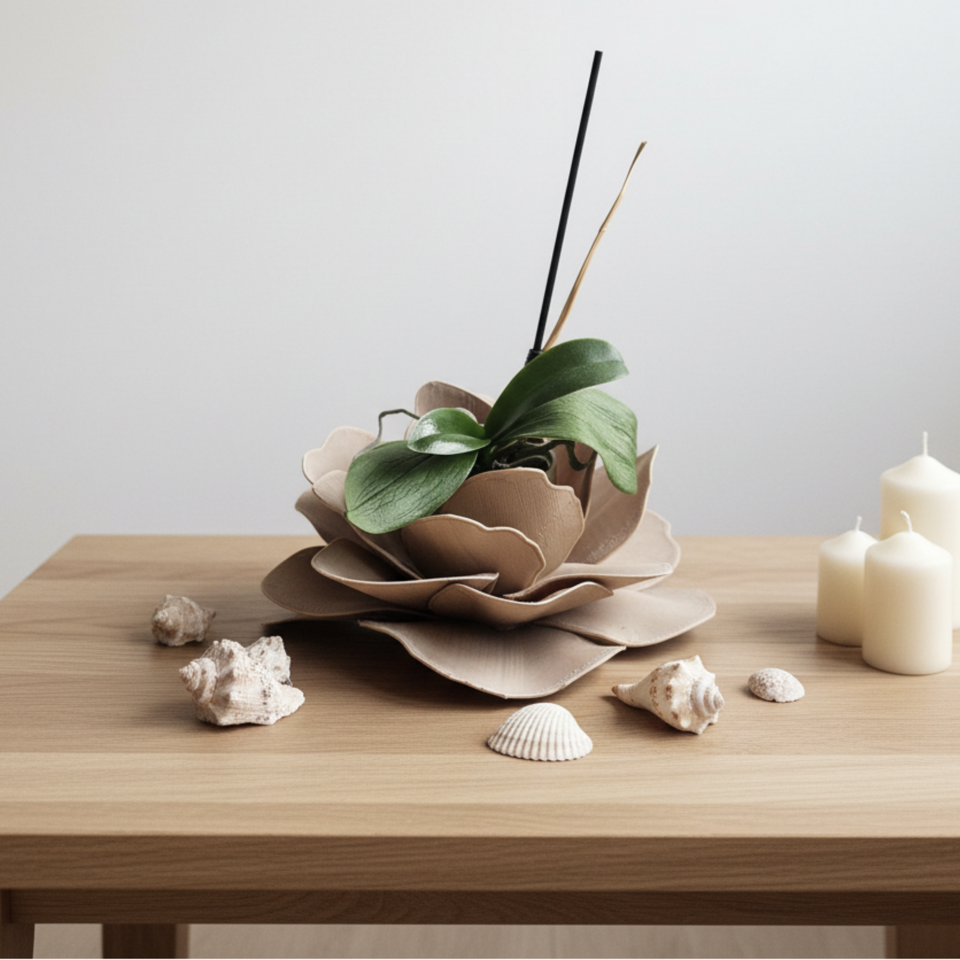 A minimalist tabletop arrangement featuring a brown flower-shaped planter holding a green leafy plant, set on a light wood table alongside white candles and small seashells against a plain light gray wall.