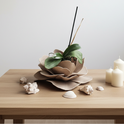 A minimalist tabletop arrangement featuring a brown flower-shaped planter holding a green leafy plant, set on a light wood table alongside white candles and small seashells against a plain light gray wall.