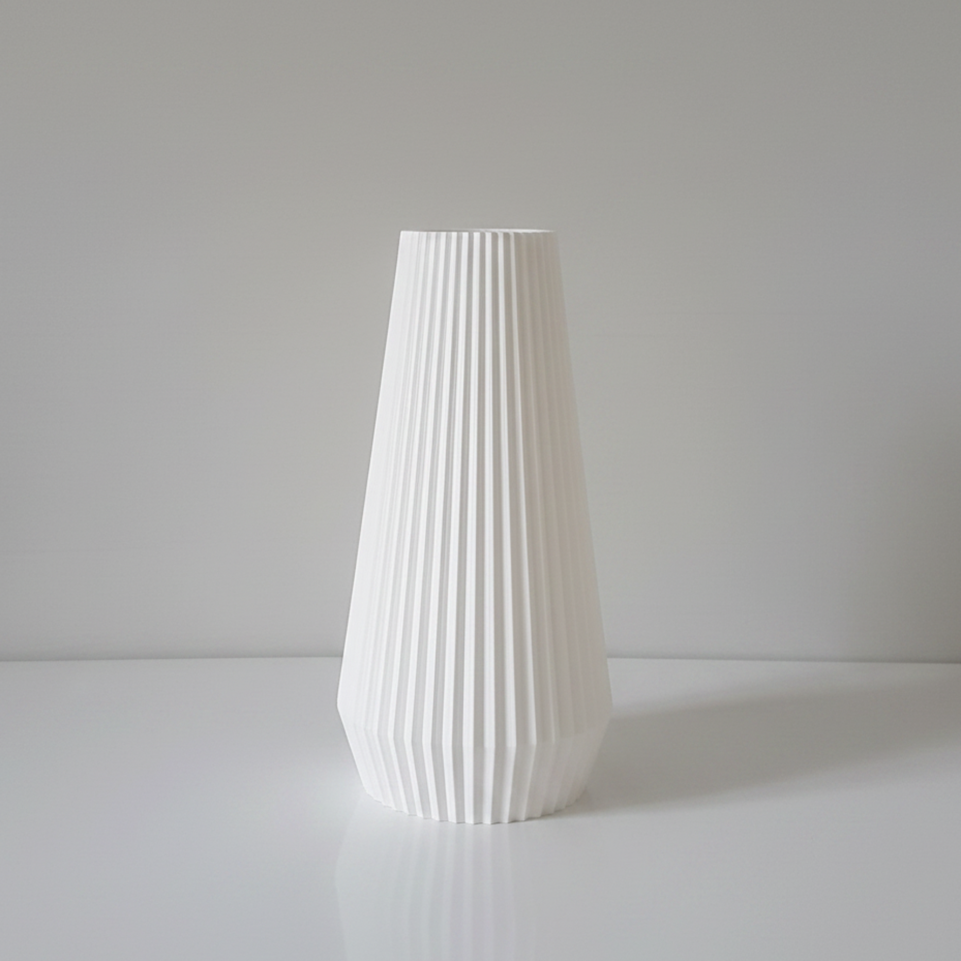 A white vase with a fluted, pleated texture, standing on a white surface against a neutral light gray background.