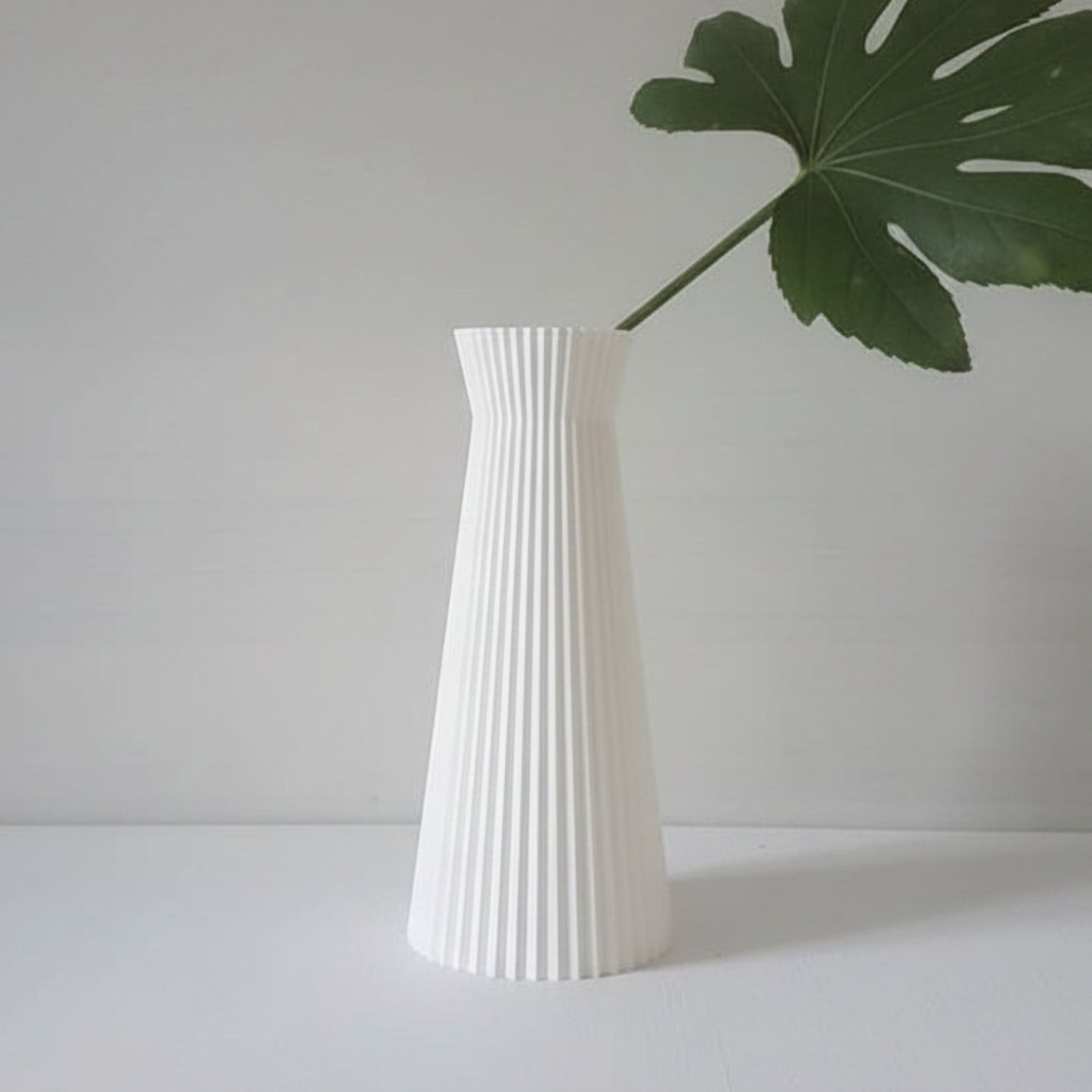 A white ribbed vase with a flared top, holding a single green monstera leaf, set against a minimalist light gray background.