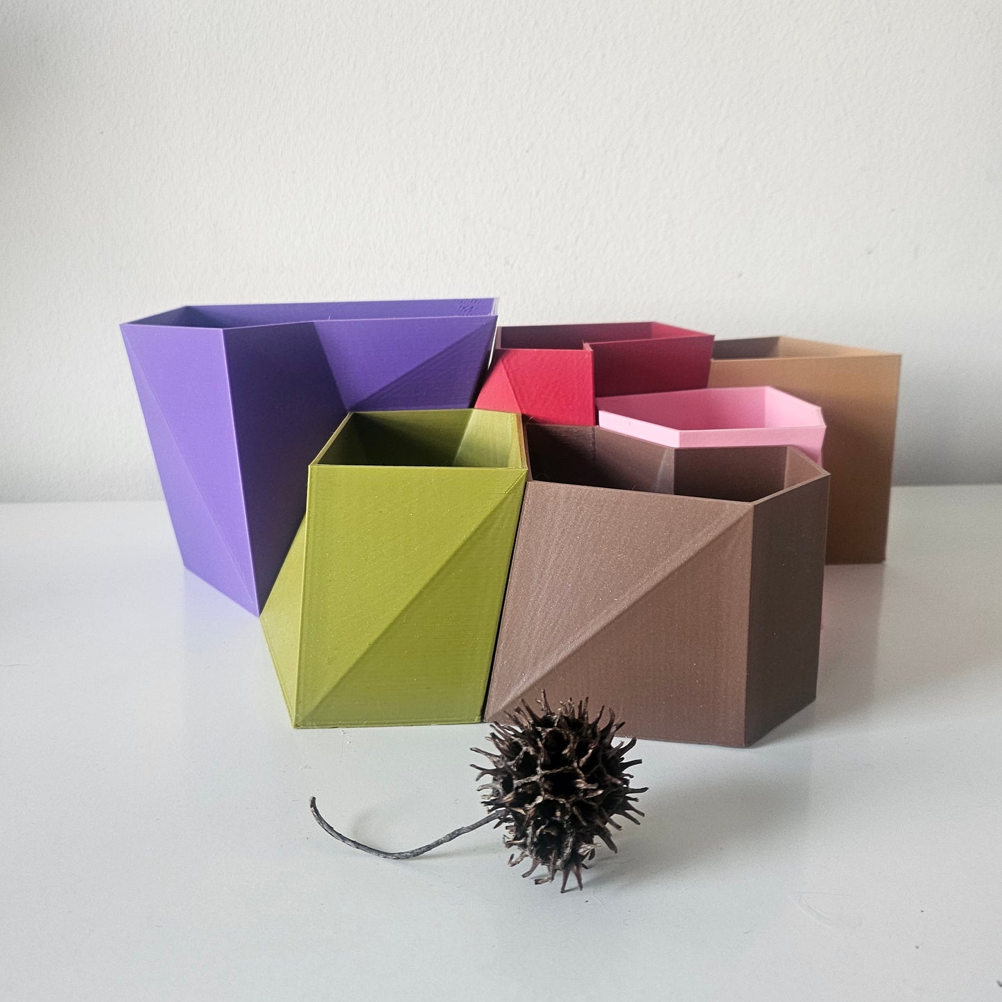 A set of geometric, container/plant pot in various colors including purple, olive green, brown, pink, and red, arranged on a white surface against a white wall, with a small dried seed pod in the foreground.