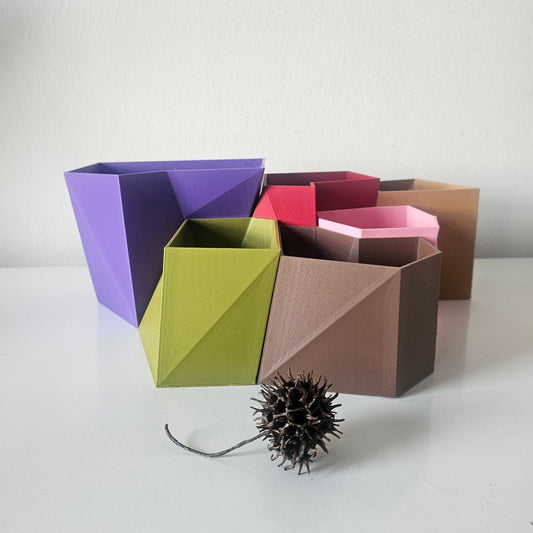 A set of geometric, container/plant pot in various colors including purple, olive green, brown, pink, and red, arranged on a white surface against a white wall, with a small dried seed pod in the foreground.