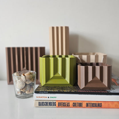 Geometric vases and planters on a shelf with books and a glass container.